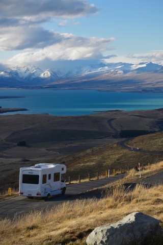 Autocaravana branca numa estrada com vista para um lago turquesa e montanhas nevadas
