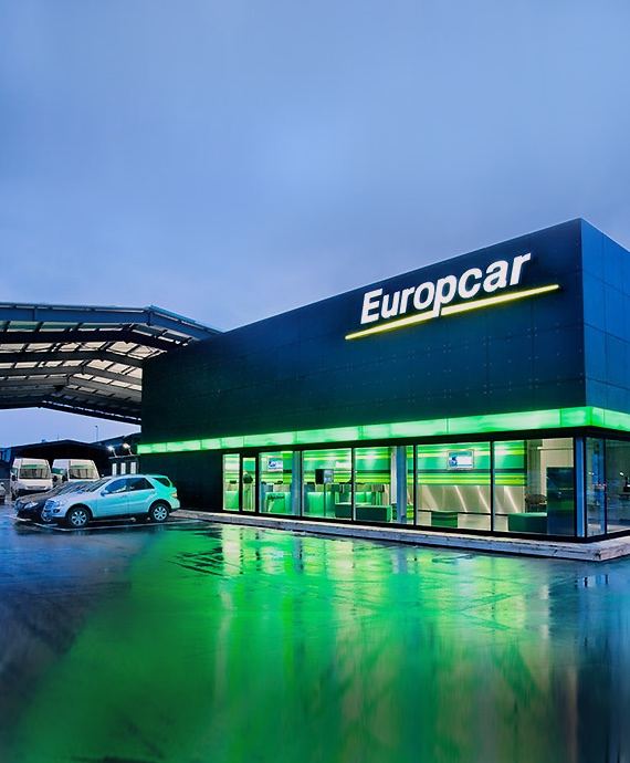 Sucursal de Europcar iluminada en verde al anochecer con coches de alquiler delante